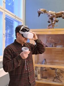 Mies testaa virtuaalitodellisuuslaseja kädessään ohjain. Taustalla vitriinissä täytettyjä lintuja.