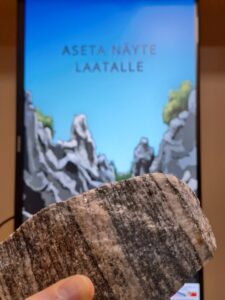 Etualalla raidallinen kivi, taustalla näyttö, jossa lukee ”Aseta näyte laatalle”.
